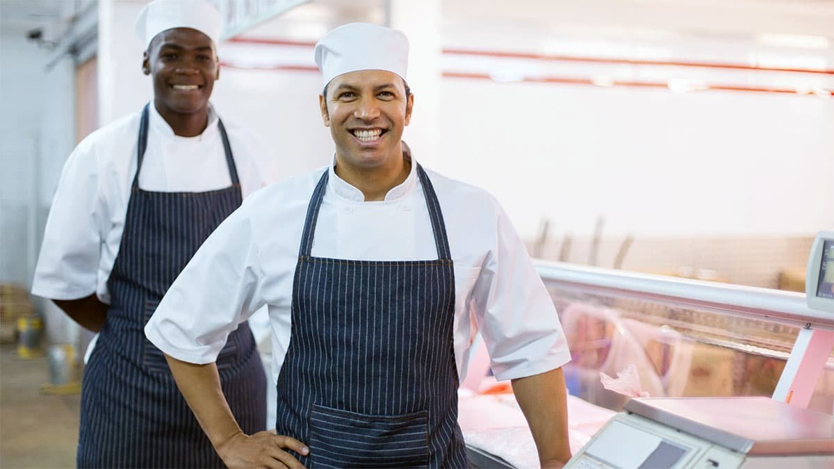 Dos empleados sonrientes con uniformes blancos y delantales, junto a una balanza